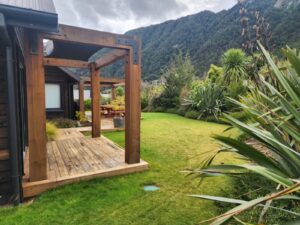 Lawn and planted bund with native plants a year after earthworks at a house in Bob’s Cove, Queenstown, with a timber pergola and bush-covered hills beyond.